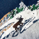 Close-up of a knit sock showing a winter landscape pattern with a brown reindeer on a snowy white background.
