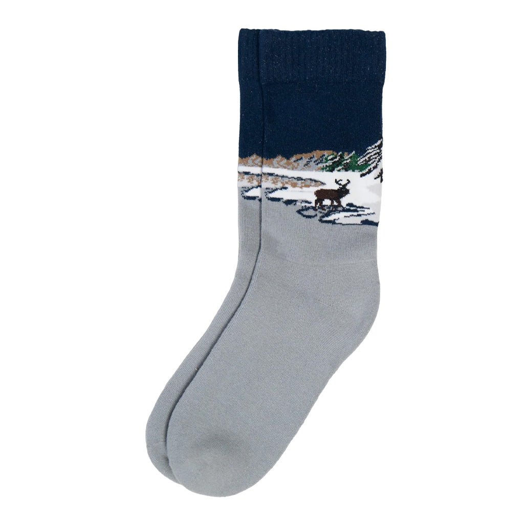 A single crew sock with a light gray foot and a dark navy blue cuff, featuring a knitted winter landscape design with a reindeer on a snowy path.
