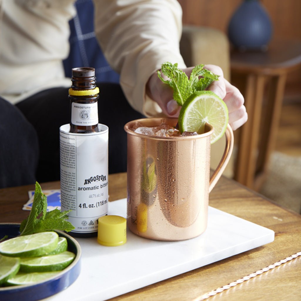 Person preparing a cocktail with a copper mug, lime, and Angostura bitters bottle on a wooden table.