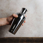 Hand holding a shiny metal cocktail shaker against a neutral background