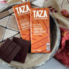 Two TAZA chocolate bars with toffee flavor next to packaging on a stone surface.