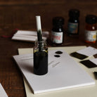 Ink bottle with a dropper on a piece of paper with writing, surrounded by ink bottles and a wooden surface.