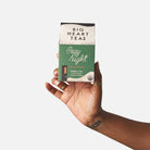 A hand holding a box of Big Heart Tea Cozy Night herbal tea against a plain light gray background.