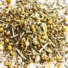 A close-up of loose-leaf chamomile tea blend, featuring dried green leaves, pale stems, and small yellow chamomile flowers scattered on a light background.