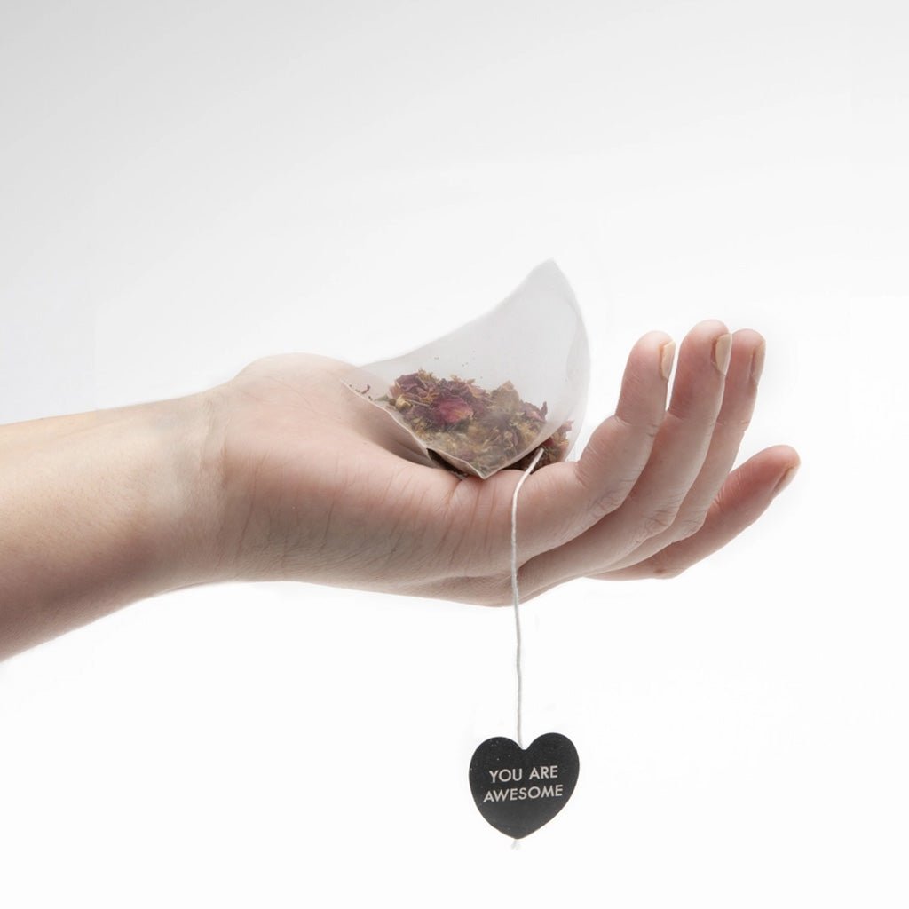 A hand holding a tea bag filled with dried tea leaves, with a heart-shaped tag hanging from the string that reads 'YOU ARE AWESOME' against a clean white background.