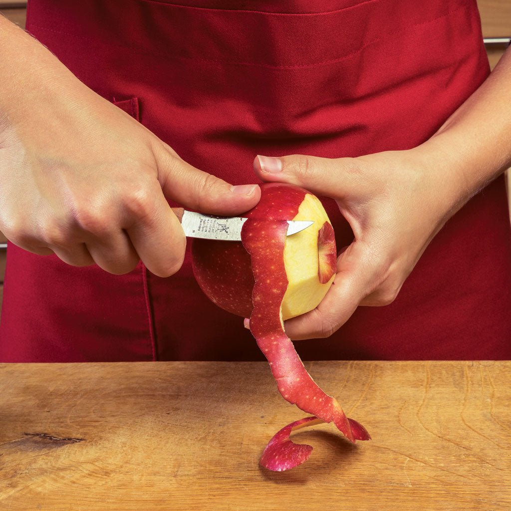 Person peeling an apple with a birds beak paring knife on a wooden surface