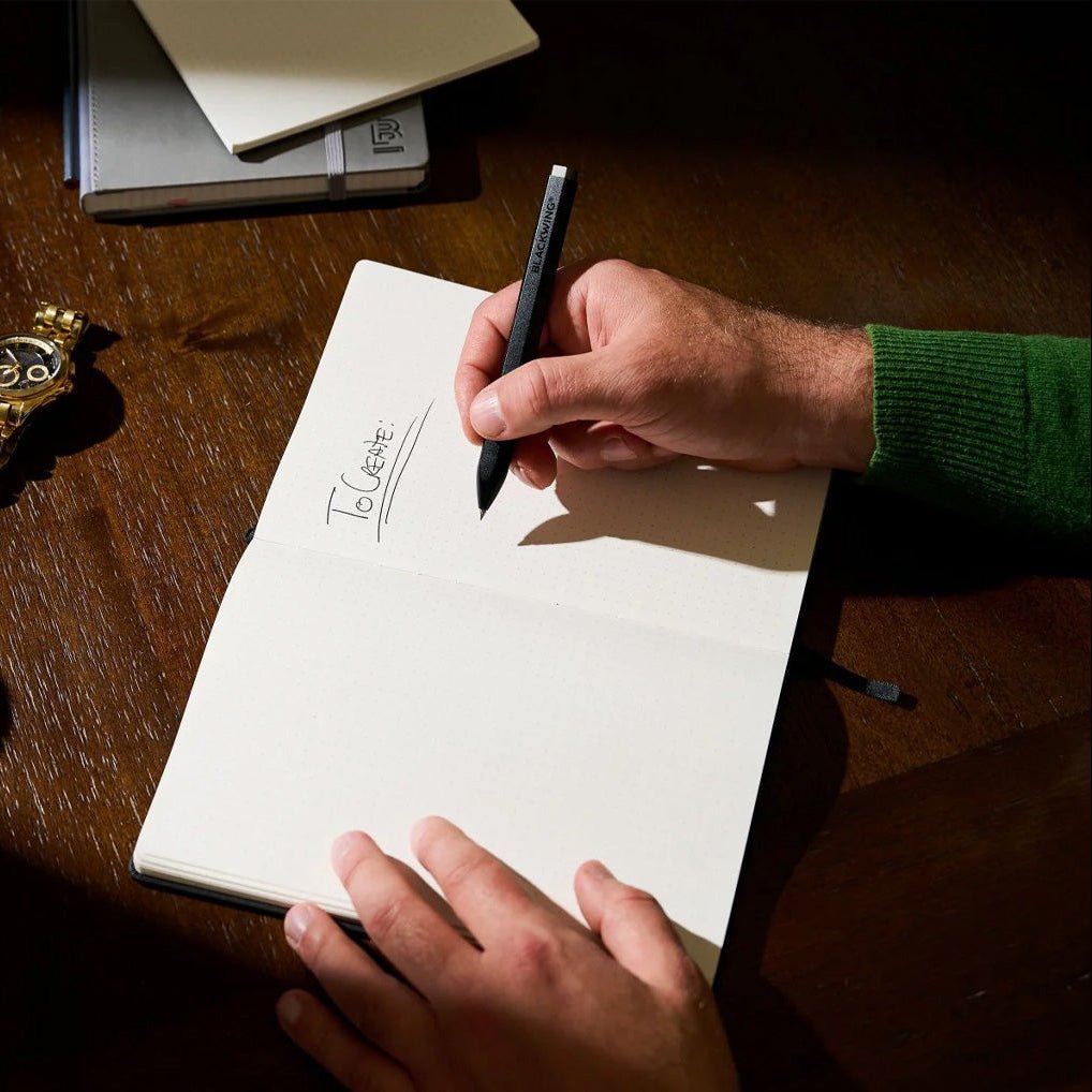 Person writing in a notebook with a Blackwing pen on a wooden surface