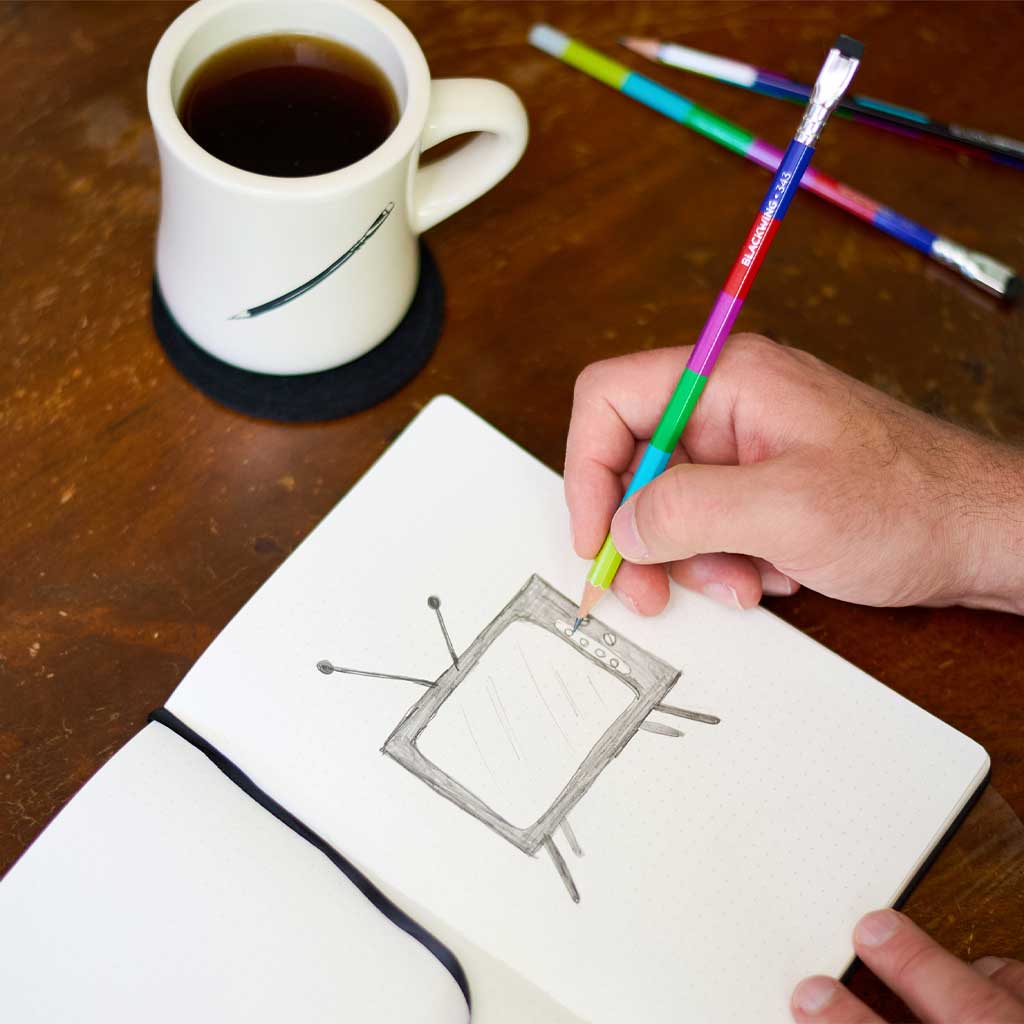 Person drawing in a notebook with colorful Blackwing 343 pencils next to a cup of coffee on a wooden table.