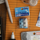 Kokuyo Kadokeshi erasers in blue and white packaging, surrounded by correction tools and a pen on an orange folder.