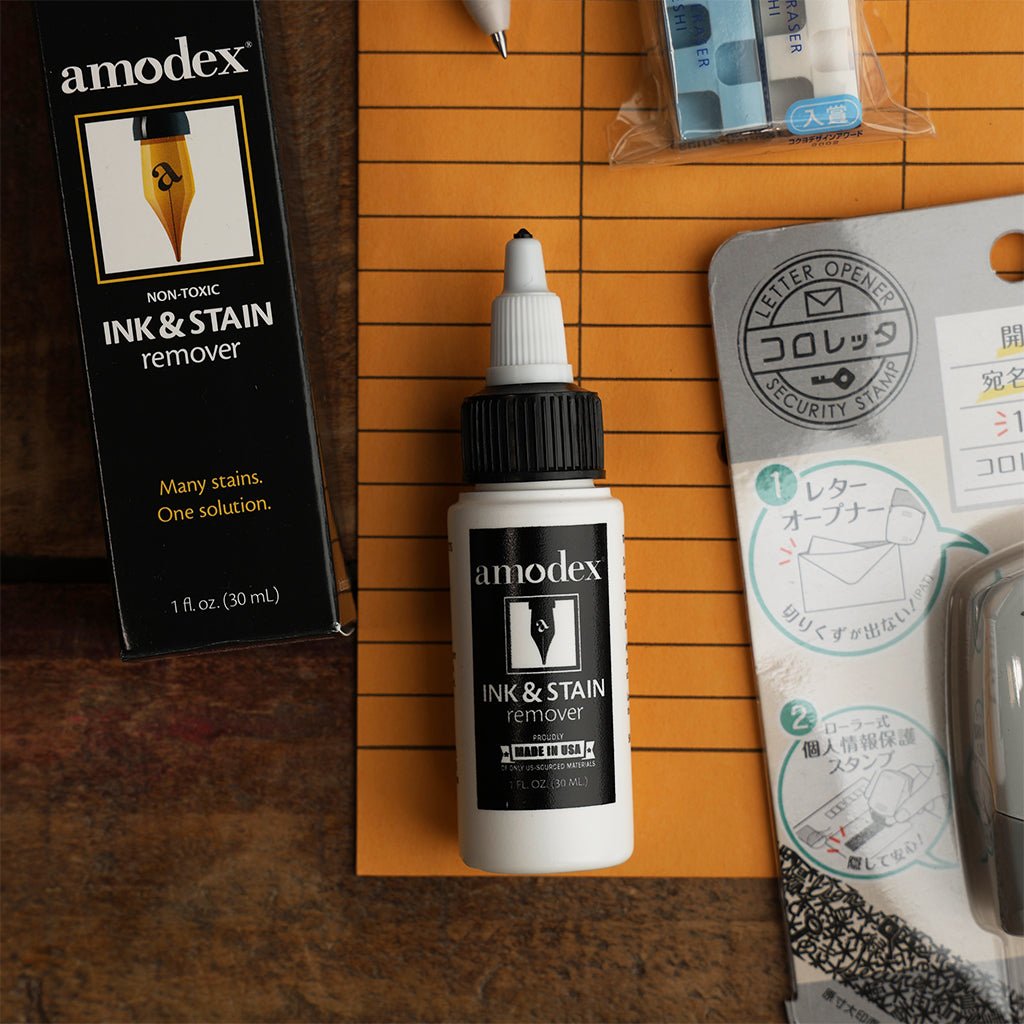Amodex ink and stain remover bottle and box beside other stationery supplies, arranged on an orange paper folder.