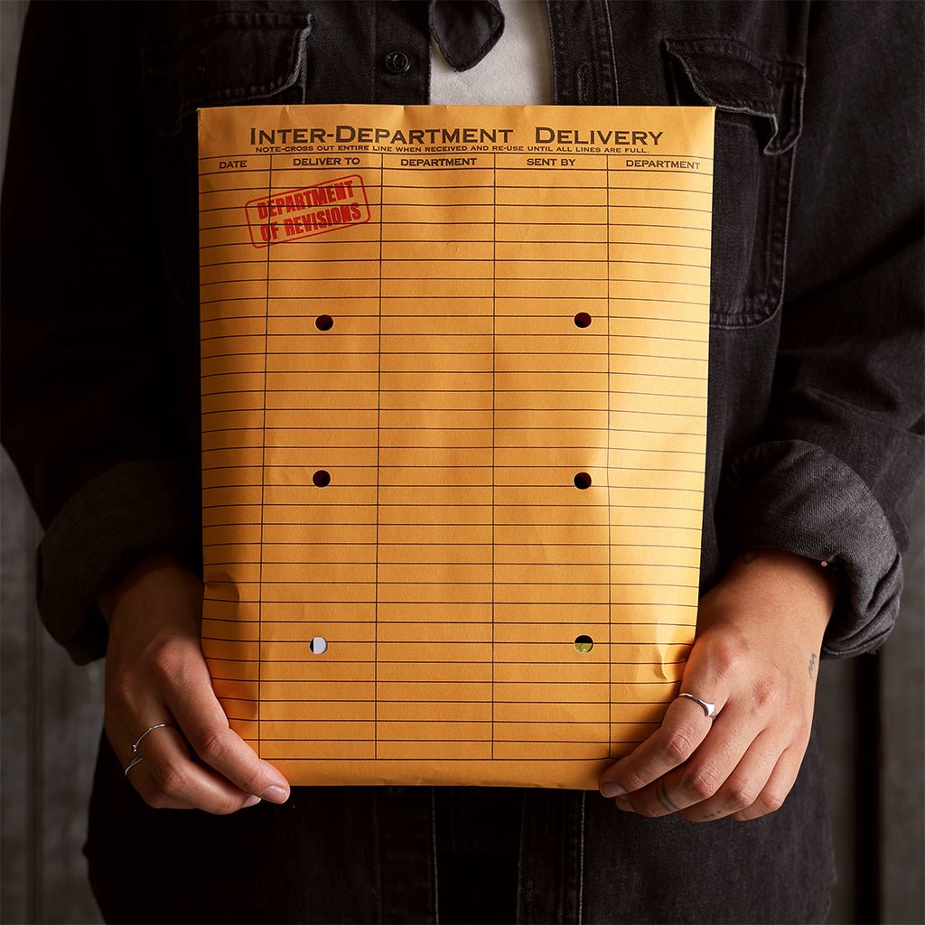 Person wearing a dark denim shirt holds an orange inter-department delivery envelope stamped “Department of Revisions.”