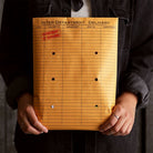 Person wearing a dark denim shirt holds an orange inter-department delivery envelope stamped “Department of Revisions.”