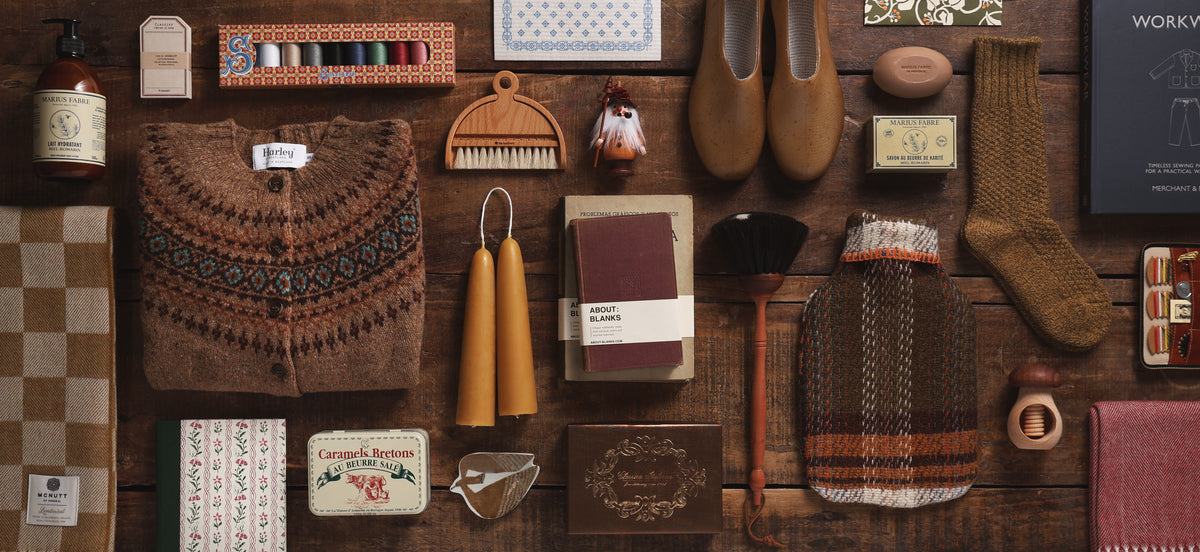 Fall essentials arranged on dark wood, including a patterned sweater, wool socks, candles, shoes, books, and home goods.
