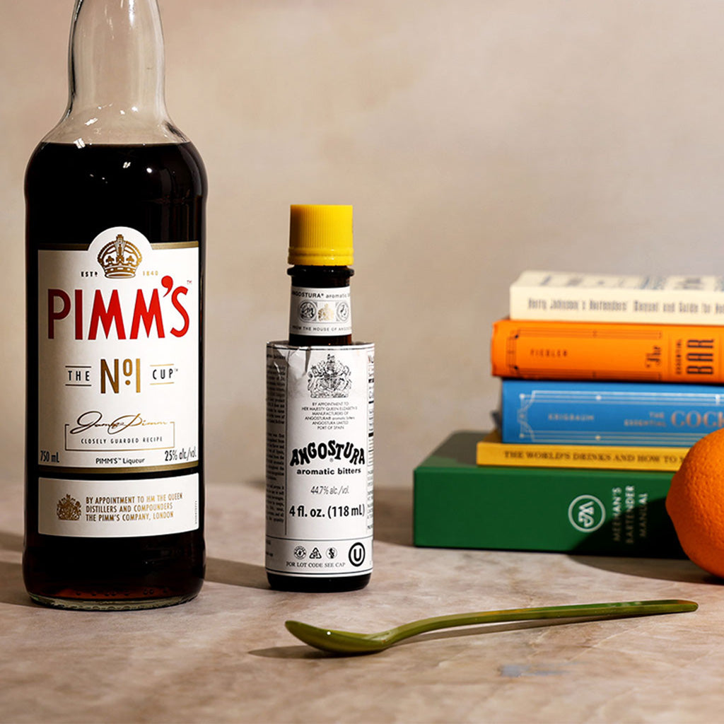 Pimm's bottle, Angostura bitters, and a green spoon on a surface with books in the background.