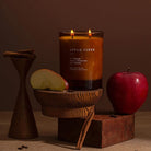 A lit double-wick candle in a clear glass jar labeled 'APPLE CIDER' sits on a wooden stand, styled with a sliced apple, a whole red apple, cinnamon sticks, and dark wood blocks against a warm brown background.