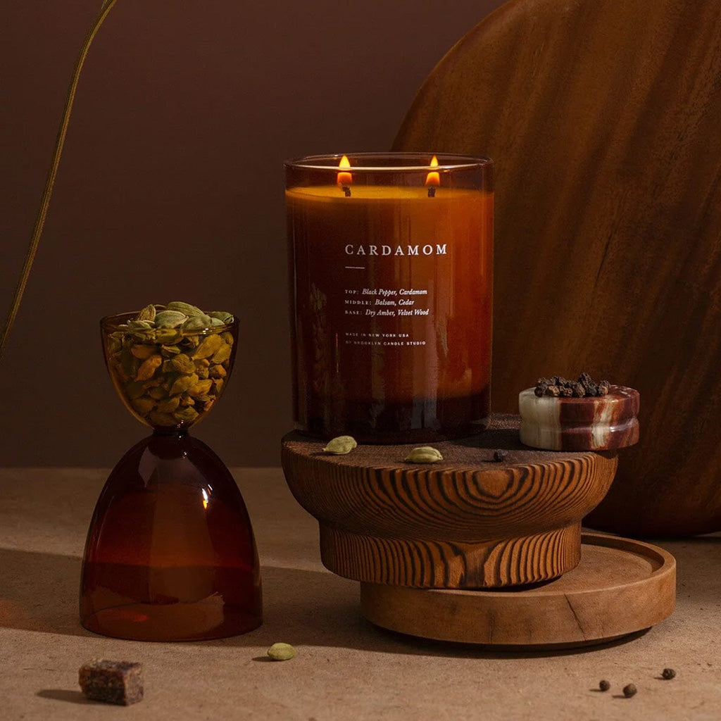 A lit double-wick candle in a clear amber glass jar labeled CARDAMOM, styled on a wooden pedestal with spices and a large wooden vessel in a warm brown setting.