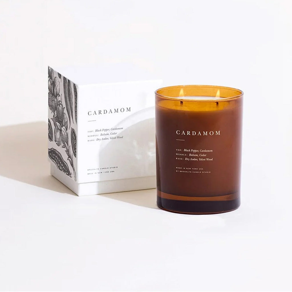 An amber glass scented candle with two wicks stands next to its white packaging box, both labeled 'CARDAMOM', on a clean white background.