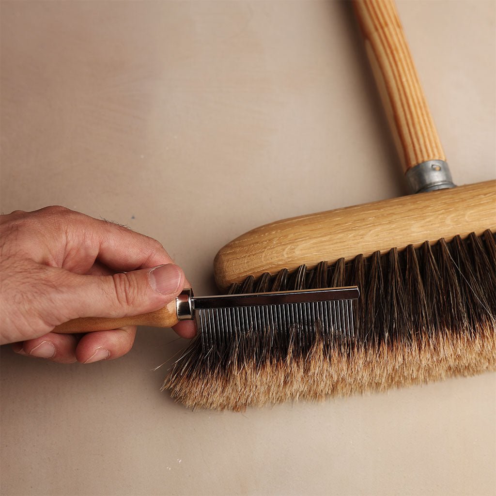 A hand holding a metal-bristle comb. The comb is being used to remove debris from broom bristles.