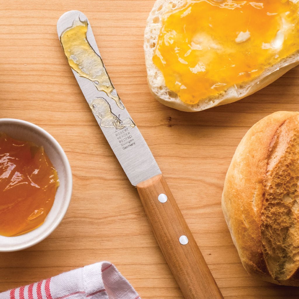 Butter knife with spreadable butter, a bowl of jam, and a piece of bread on a wooden surface.