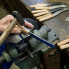 Hand holding a hammer over a blade being inserted into a wooden handle with tools on a workbench