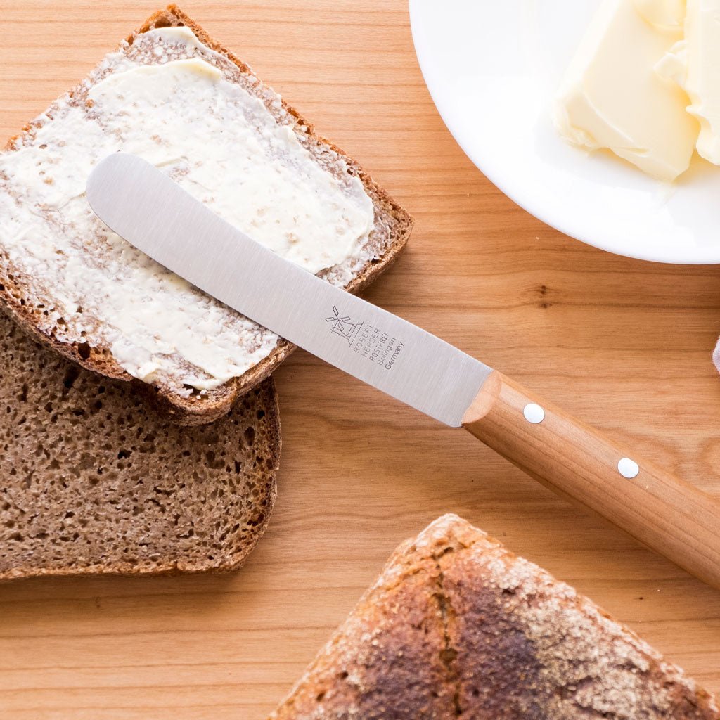Breakfast knife spreading butter on a slice of bread with more bread and butter in the background on a wooden surface.