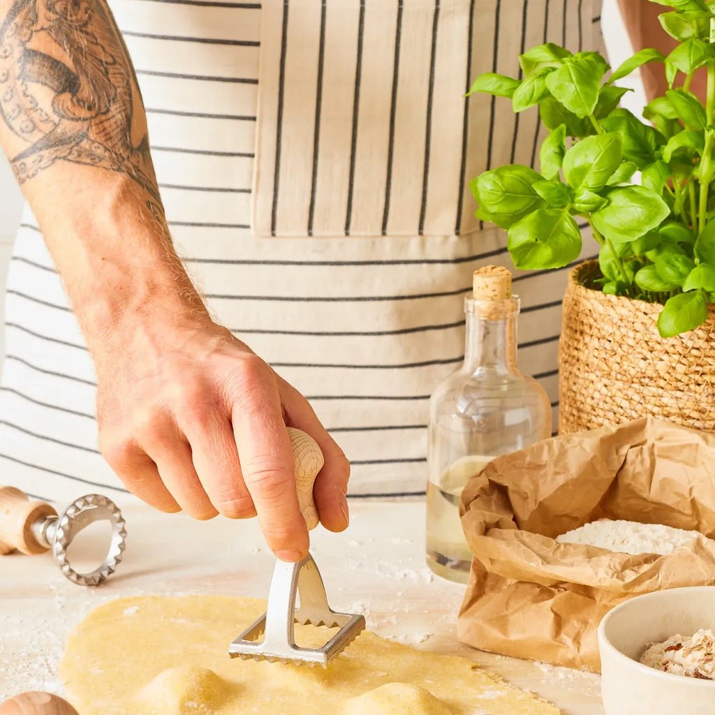A close-up lifestyle shot of a person wearing a striped apron using a metal pastry cutter with a wooden handle to cut dough on a floured surface, surrounded by baking ingredients and a potted basil plant.