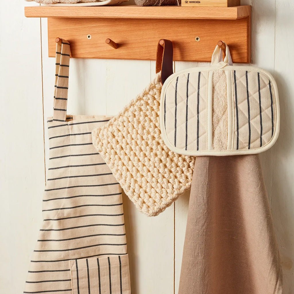 A wooden wall-mounted hook rack with three hanging kitchen accessories: a beige striped apron, a chunky knit cream pot holder, and a quilted cream pot holder with dark stripes, against a white paneled wall.