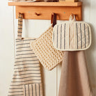 A wooden wall-mounted hook rack with three hanging kitchen accessories: a beige striped apron, a chunky knit cream pot holder, and a quilted cream pot holder with dark stripes, against a white paneled wall.