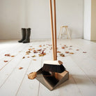 A metal dustpan and matching broom set with light wood handles, placed on a white wooden floor with scattered autumn leaves. A pair of dark rain boots and a small wooden stool are visible in the softly lit background.
