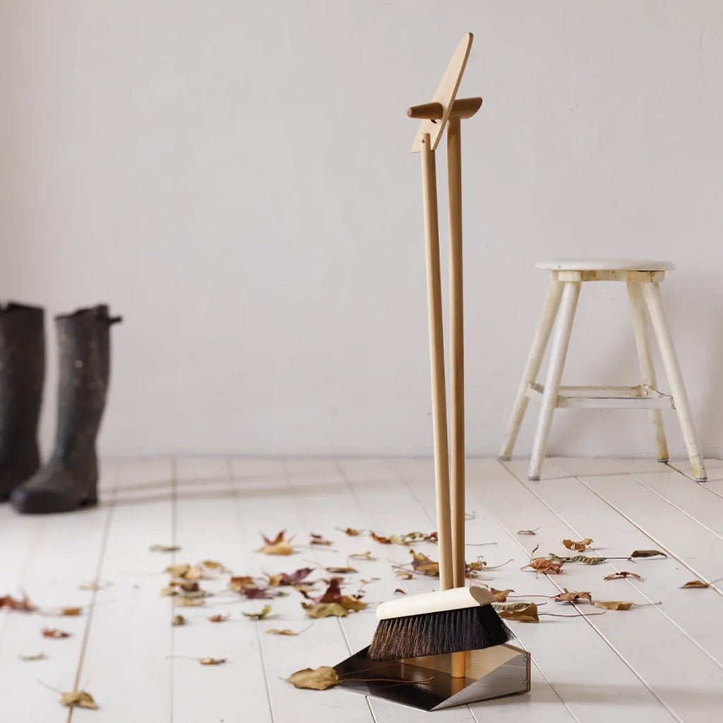 A wooden push broom with a matching metal dustpan stands on a white wooden floor scattered with dry leaves, with a white stool and dark rain boots in the background.