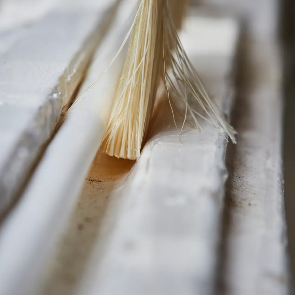 A close-up shot of a natural cleaning brush with light tan bristles, positioned inside the groove of a white textured wall or siding.