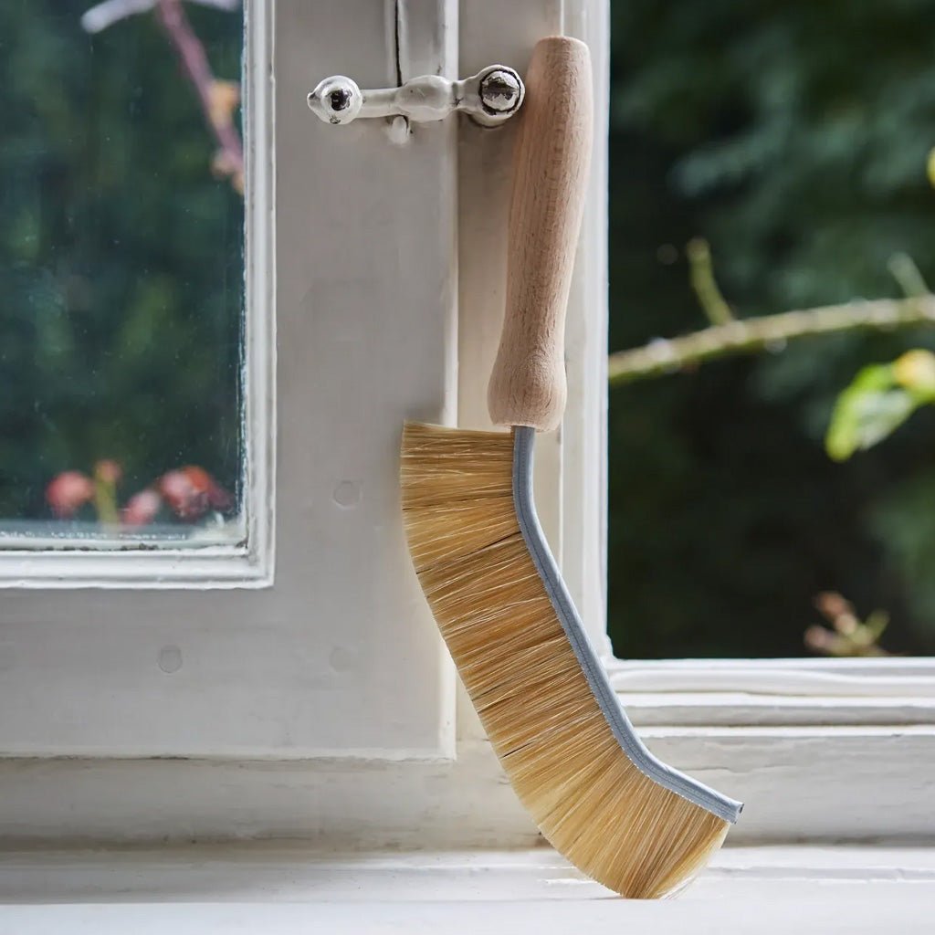 A window cleaning brush with a light wood handle and natural bristles, leaning on a white window frame with an outdoor garden background.