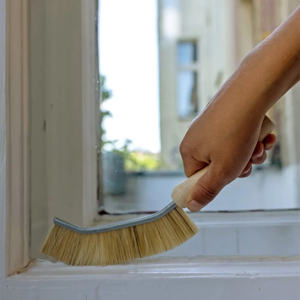 A hand holding a window cleaning brush with natural bristles and a light wood handle, cleaning a white window sill.