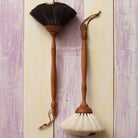 Two wooden dust brushes with long handles hang against a distressed light wood panel background. One brush has dark brown bristles and the other has off-white bristles, both with wood handles and leather ties.