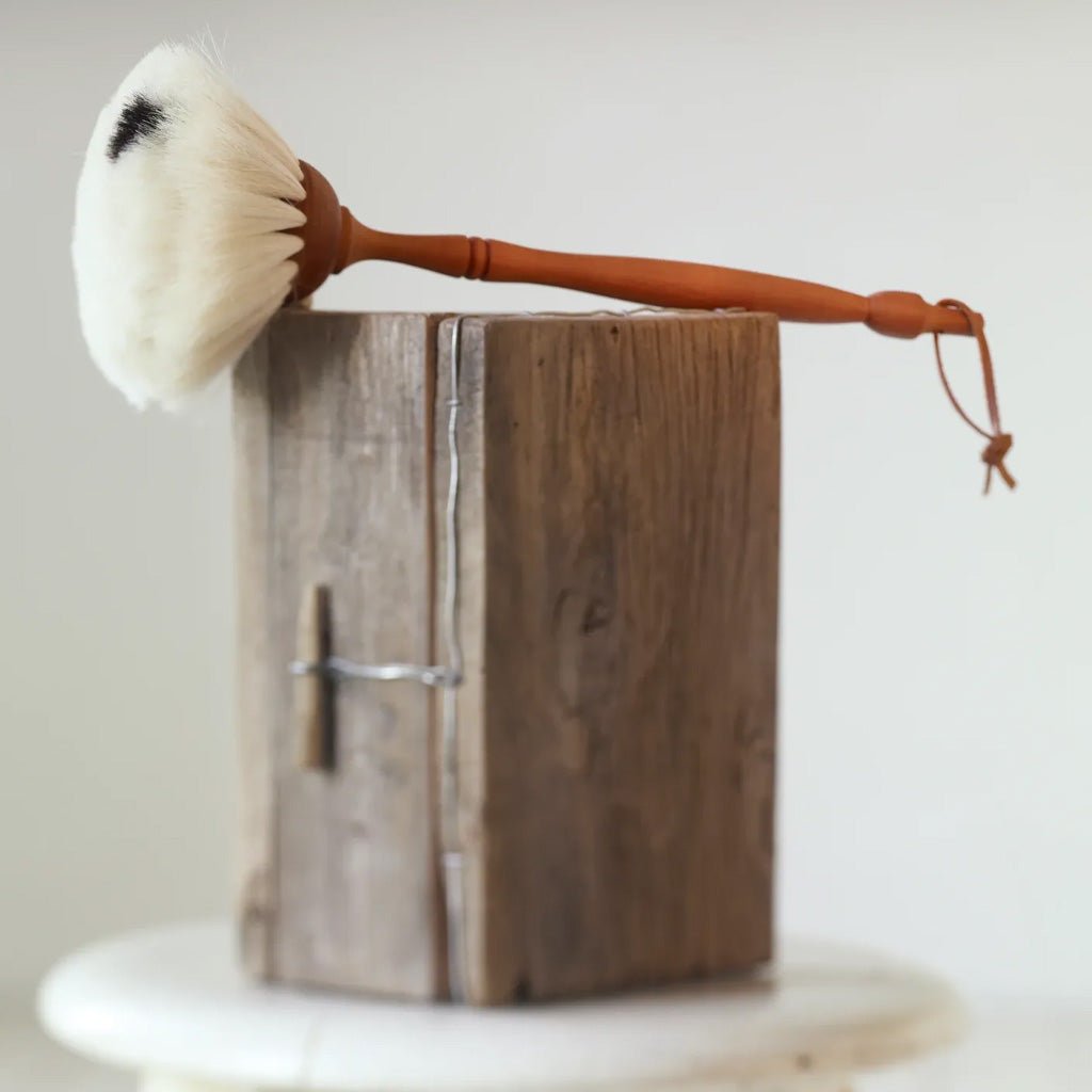 A goat hair dust brush with a light brown wooden handle and white bristles, resting on top of a rustic wooden block.