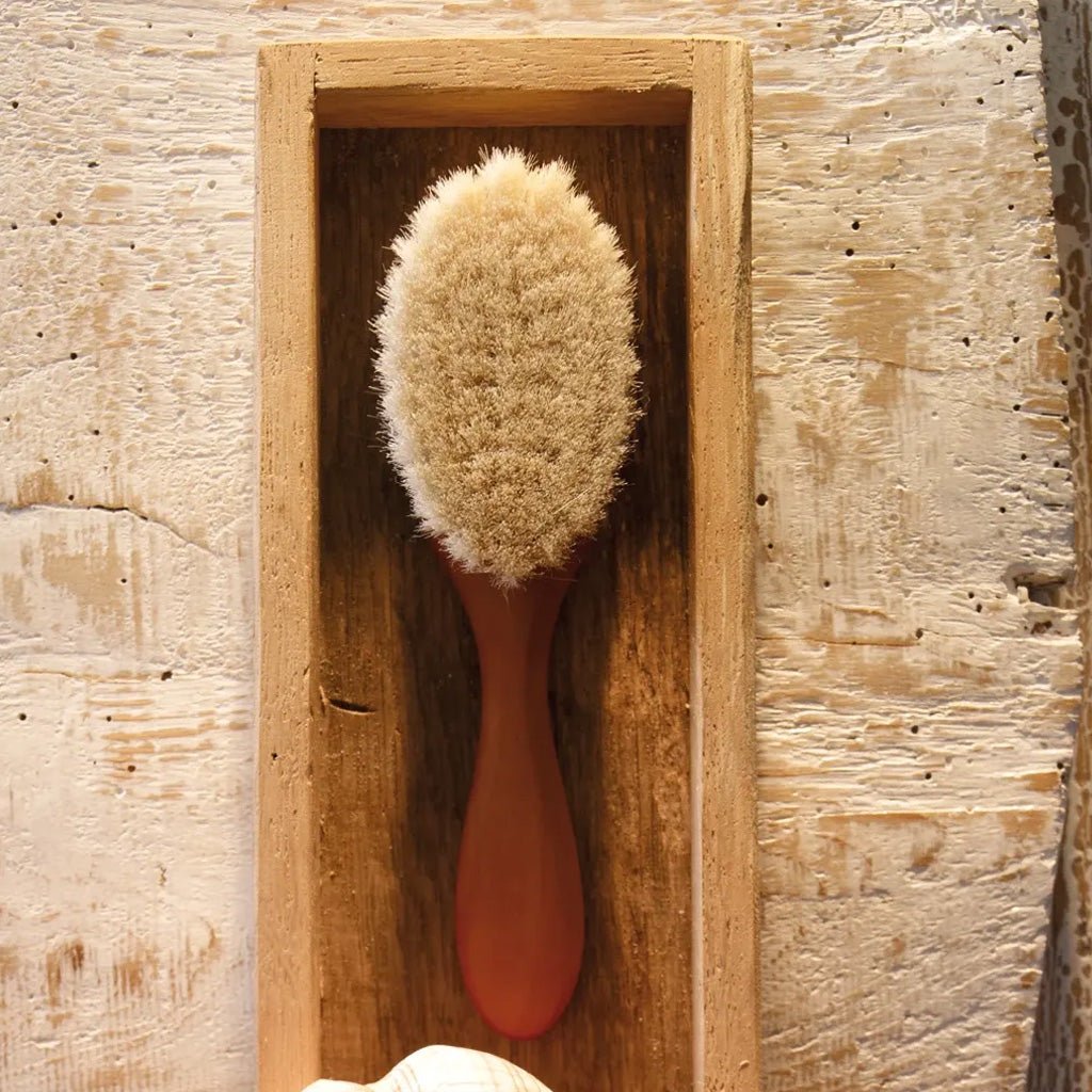 A wooden hair brush with a reddish-brown handle and natural beige bristles, displayed inside a rectangular wooden holder against a textured light wood background.