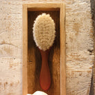 A wooden hair brush with a reddish-brown handle and natural beige bristles, displayed inside a rectangular wooden holder against a textured light wood background.