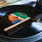 A close-up of a black vinyl record on a turntable, with a wooden cleaning brush resting across the record and a tonearm visible at the top right.