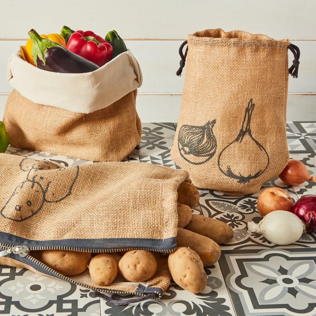 Three reusable fabric drawstring and zip pouches made from natural burlap are arranged on a patterned tile surface with vegetables and potatoes around them. The burlap has black printed illustrations of onions and garlic.