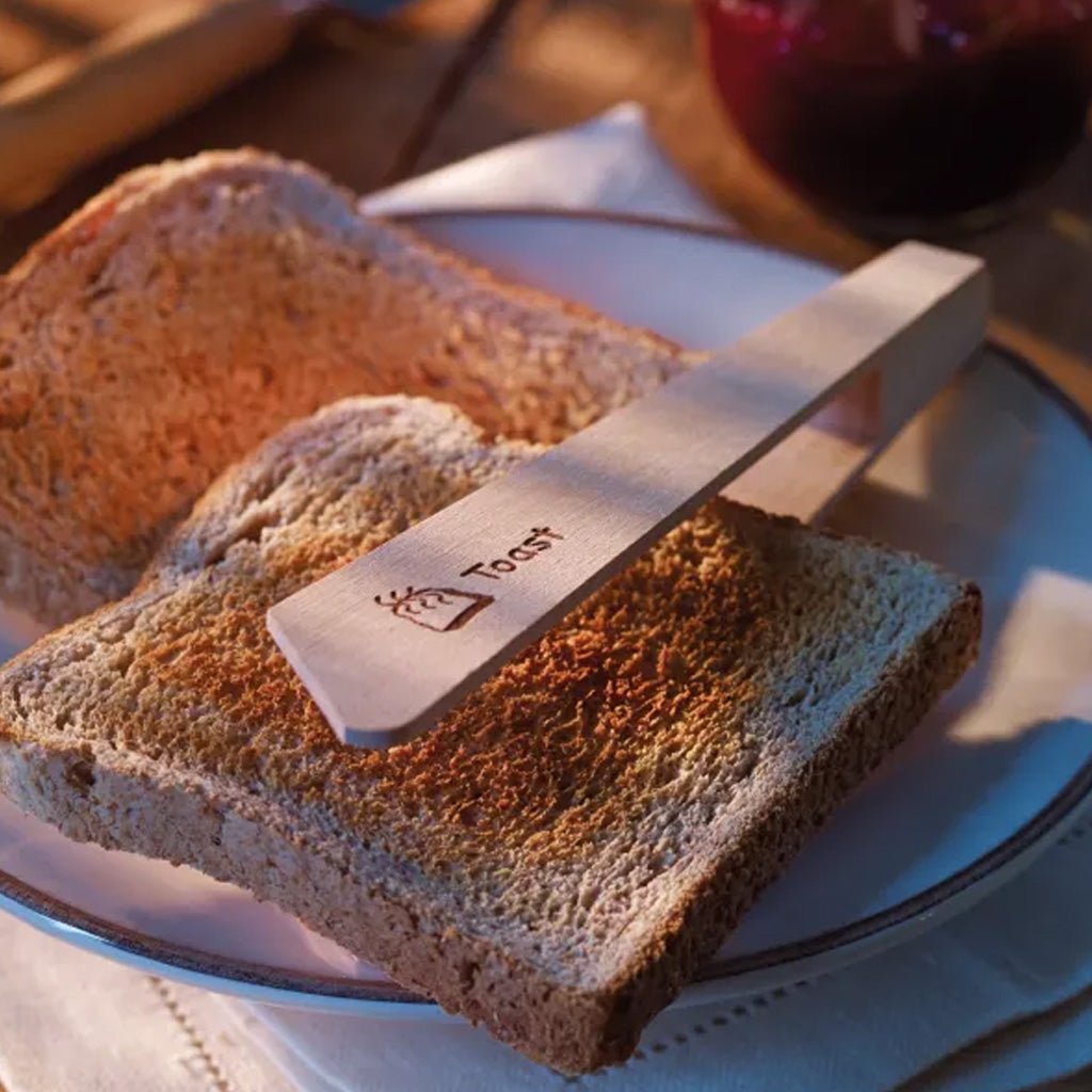 A wooden toast tong with 'Toast' engraved on it, resting on a slice of toasted bread on a white plate.
