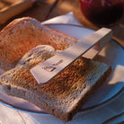 A wooden toast tong with 'Toast' engraved on it, resting on a slice of toasted bread on a white plate.