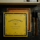 A yellow calligraphy guide sheet with black ornamental border and text is displayed in front of a matching orange book. Two calligraphy brushes with dark handles and gold ferrules lean against a dark textured surface on the right.