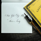 A calligraphy pen with a black and gold handle and wooden grip rests on an open white grid notebook showing elegant cursive writing, with a yellow decorative book partially visible in the corner on a dark textured surface.