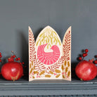 A decorative paper card with a Christmas bird and botanical pattern, styled on a gray mantelpiece with red pomegranates and berry sprigs against a dark gray wall.