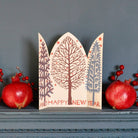 A decorative arched holiday card reading 'AND HAPPY NEW YEAR' with three stylized trees and small red stars, displayed on a gray mantel with pomegranates and red berries against a gray wall.