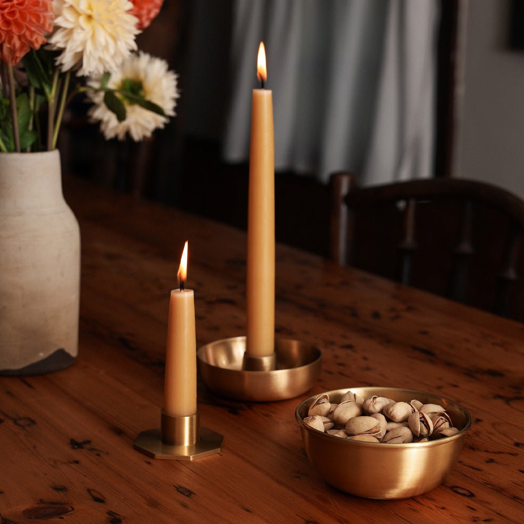 Natural taper candles lit in brass holders, next to bowl of nuts on table.