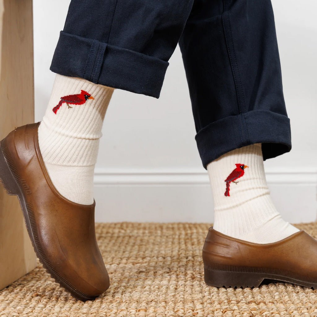 A person wearing dark navy pants, white socks with red cardinal bird embroidery, and brown clogs on a woven jute rug.