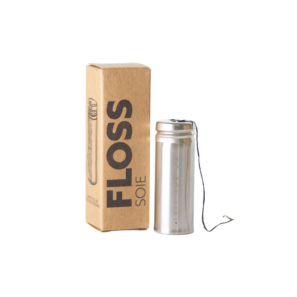 Charcoal floss in stainless steel holder next to kraft box packaging on white background