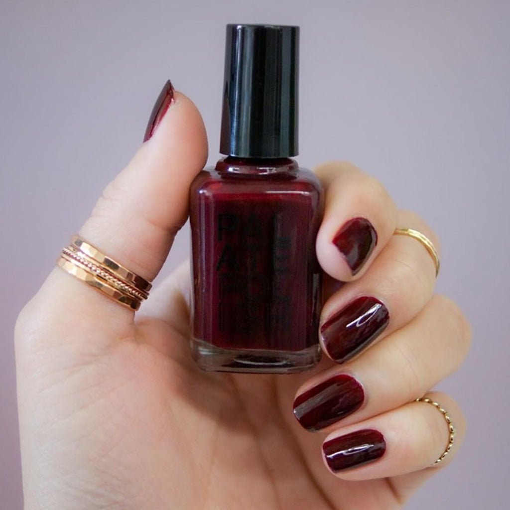 Hand holding a bottle of dark red nail polish with matching nails against a neutral background