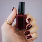 Hand holding a bottle of dark red nail polish with matching nails against a neutral background